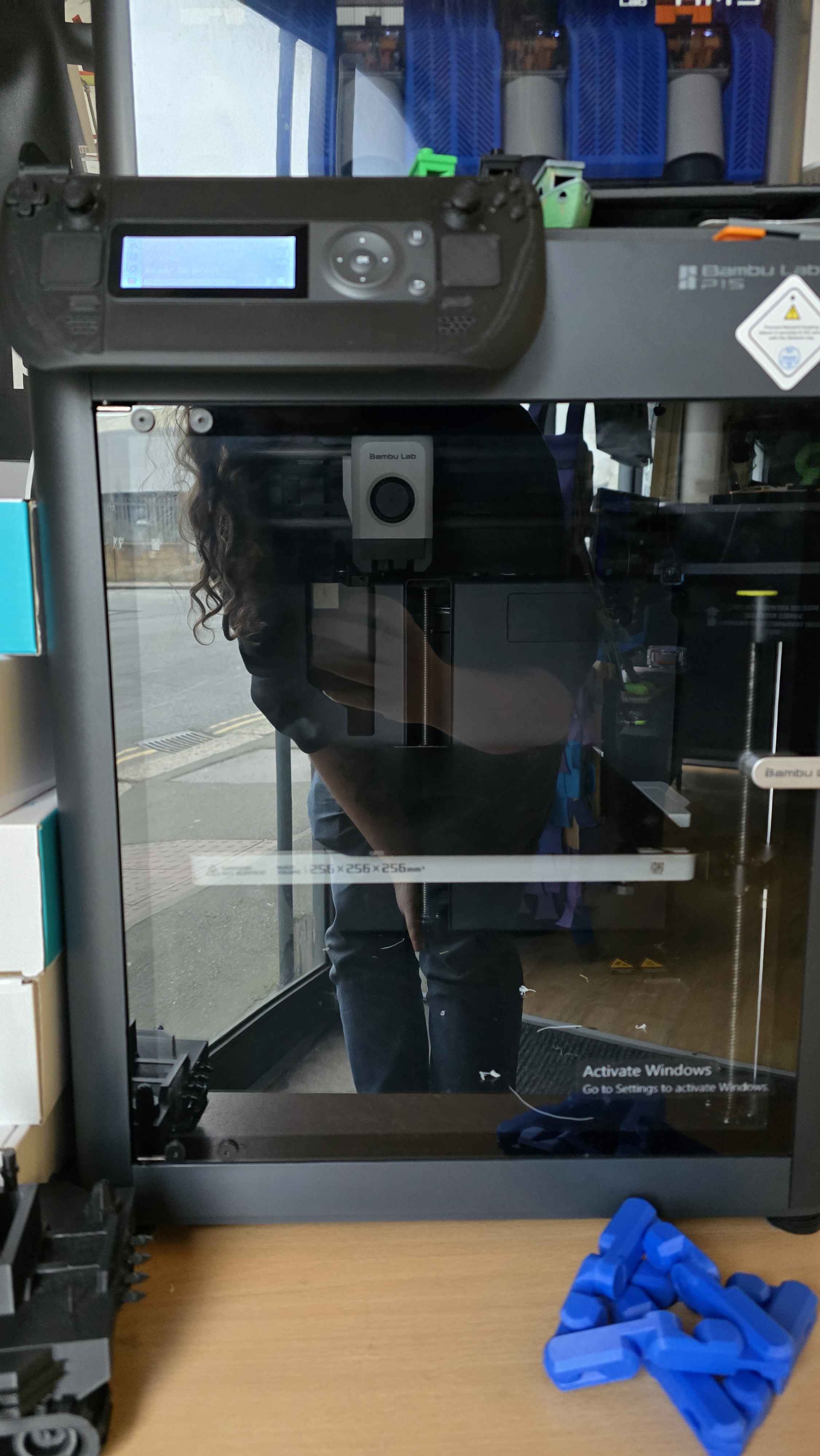 A Bambu Lab 3D printer in action, creating blue 3D printed components. The transparent enclosure allows students to observe the printing process and understand how digital designs become physical objects.
