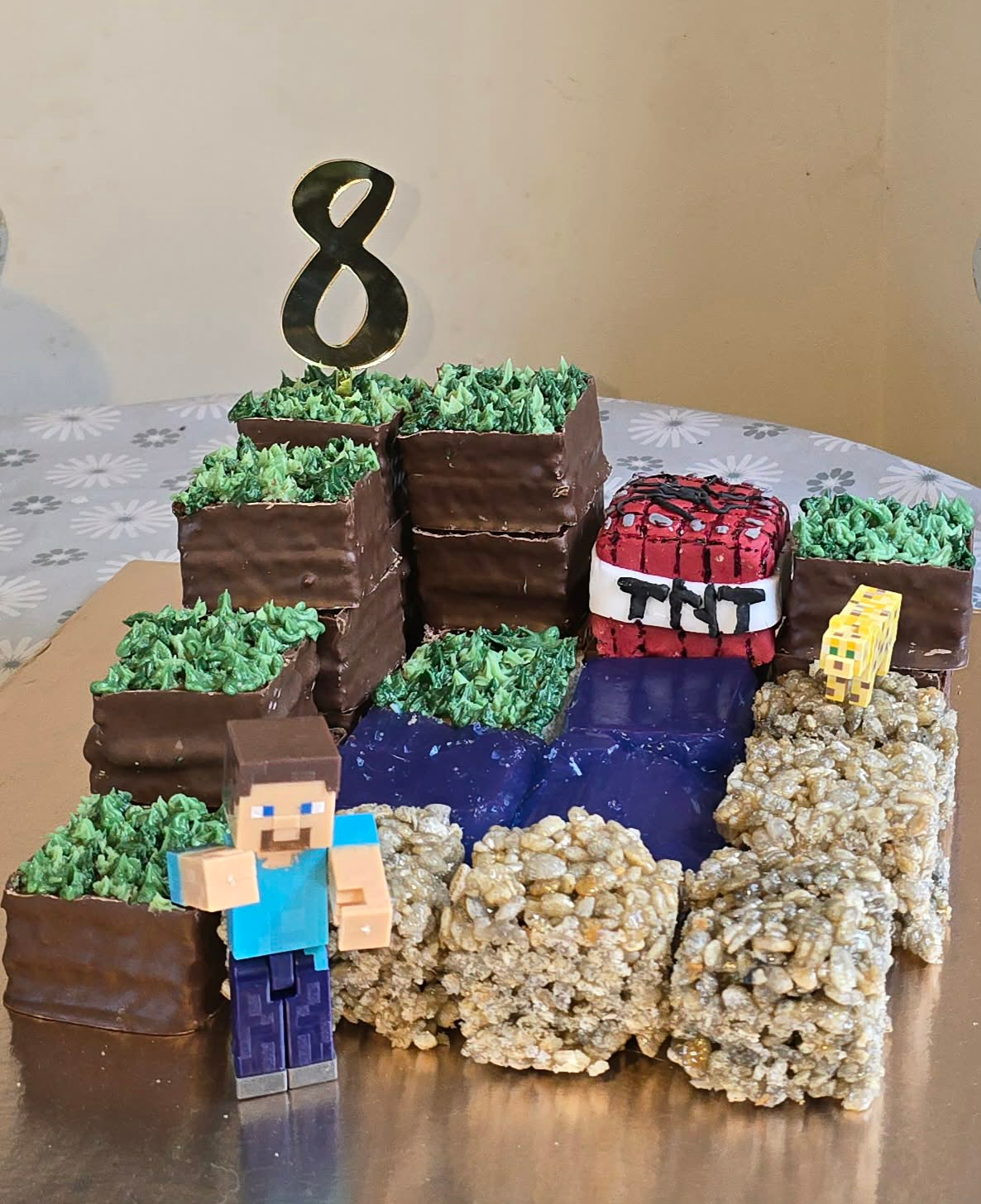An incredible Minecraft-themed birthday cake featuring chocolate blocks with green grass frosting, rice crispy treat sand blocks, and a blue water feature. Complete with a Steve character figure and topped with a number 8 candle, this creative cake perfectly captures the blocky aesthetic of the game.
